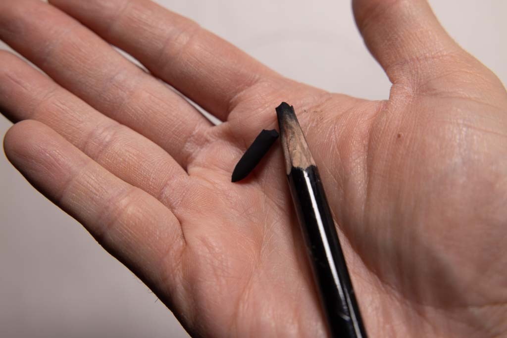 Broken black pencil core resting in a hand beside a sharpened Conté pencil