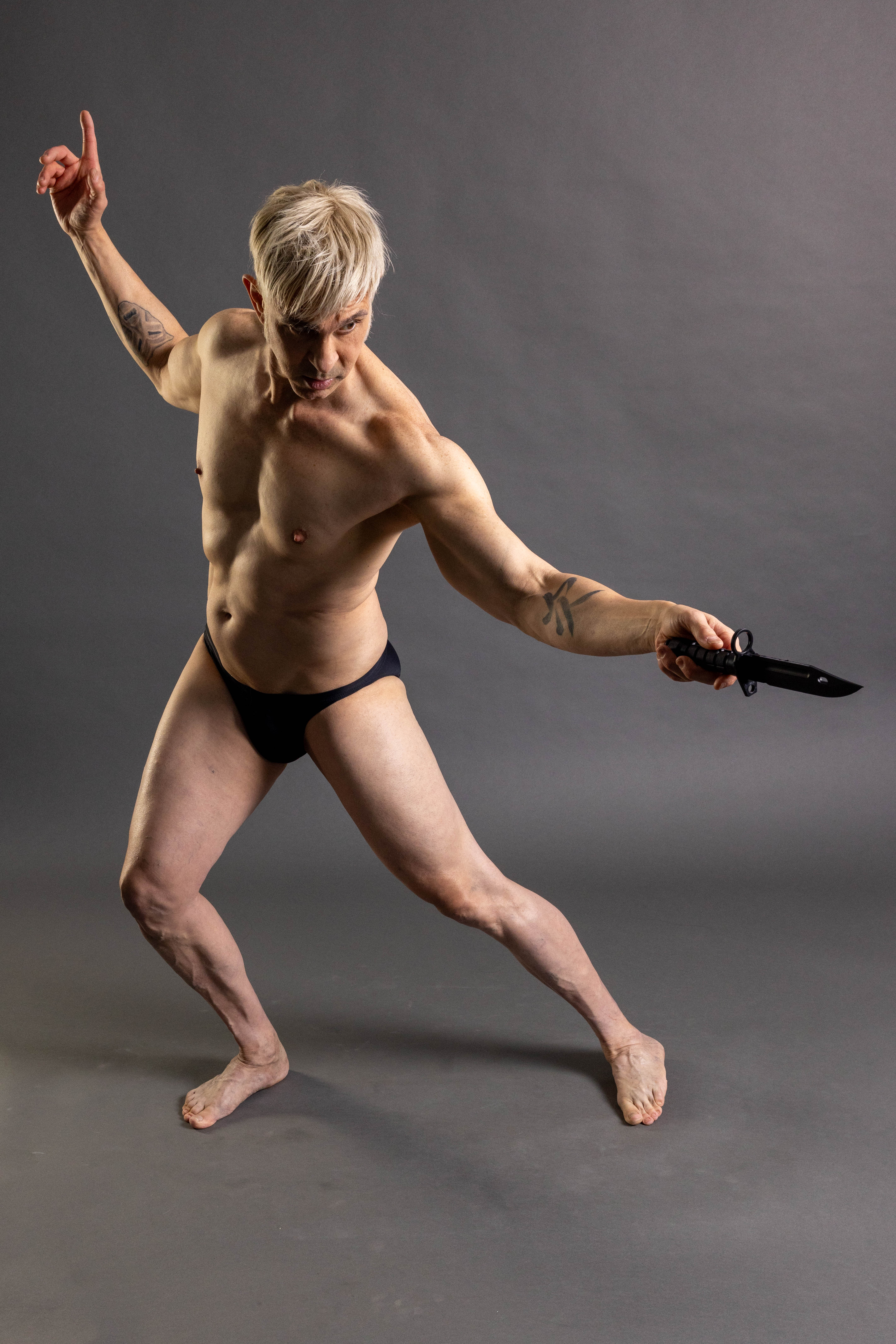 Wide standing action pose reference with one arm lifted and the other extended holding a knife prop