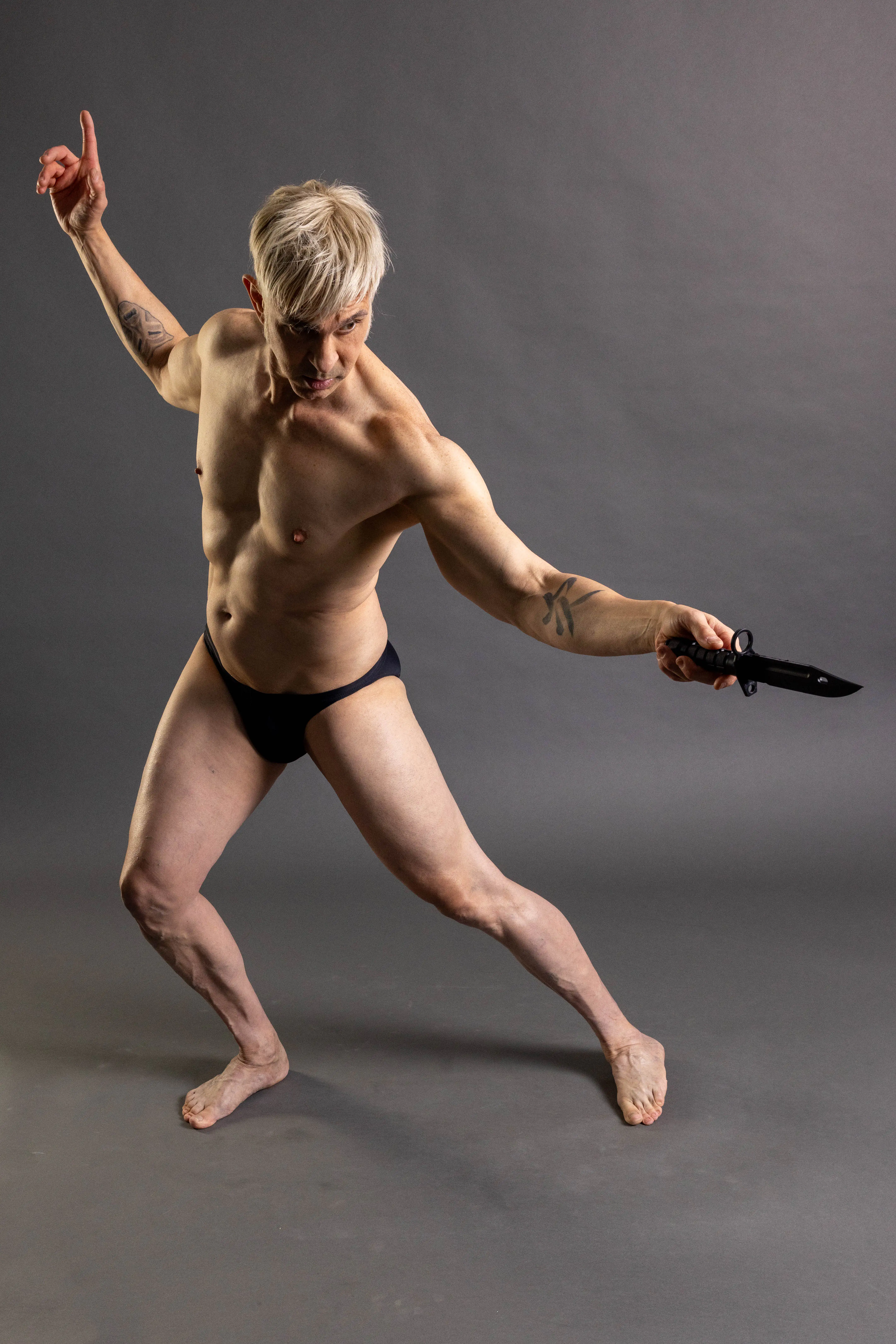 Wide standing action pose reference with one arm lifted and the other extended holding a knife prop